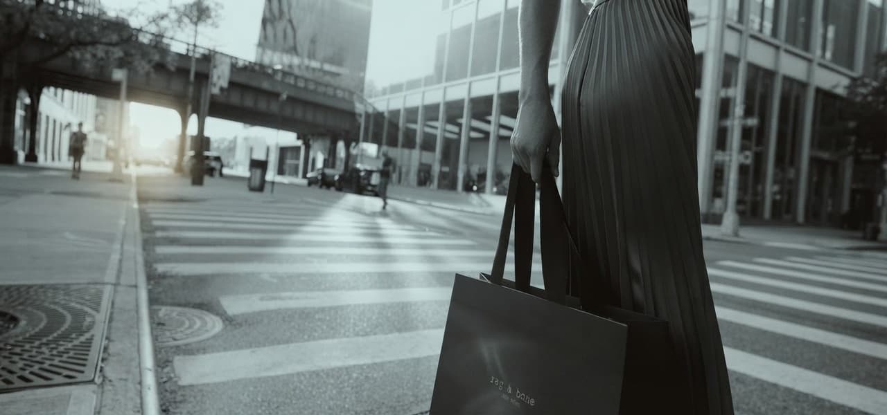 Una persona in una gonna plissettata tiene due shopping bag nere mentre si trova su una strada pedonale di una zona urbana, con edifici e un cavalcavia sullo sfondo. | Webranking